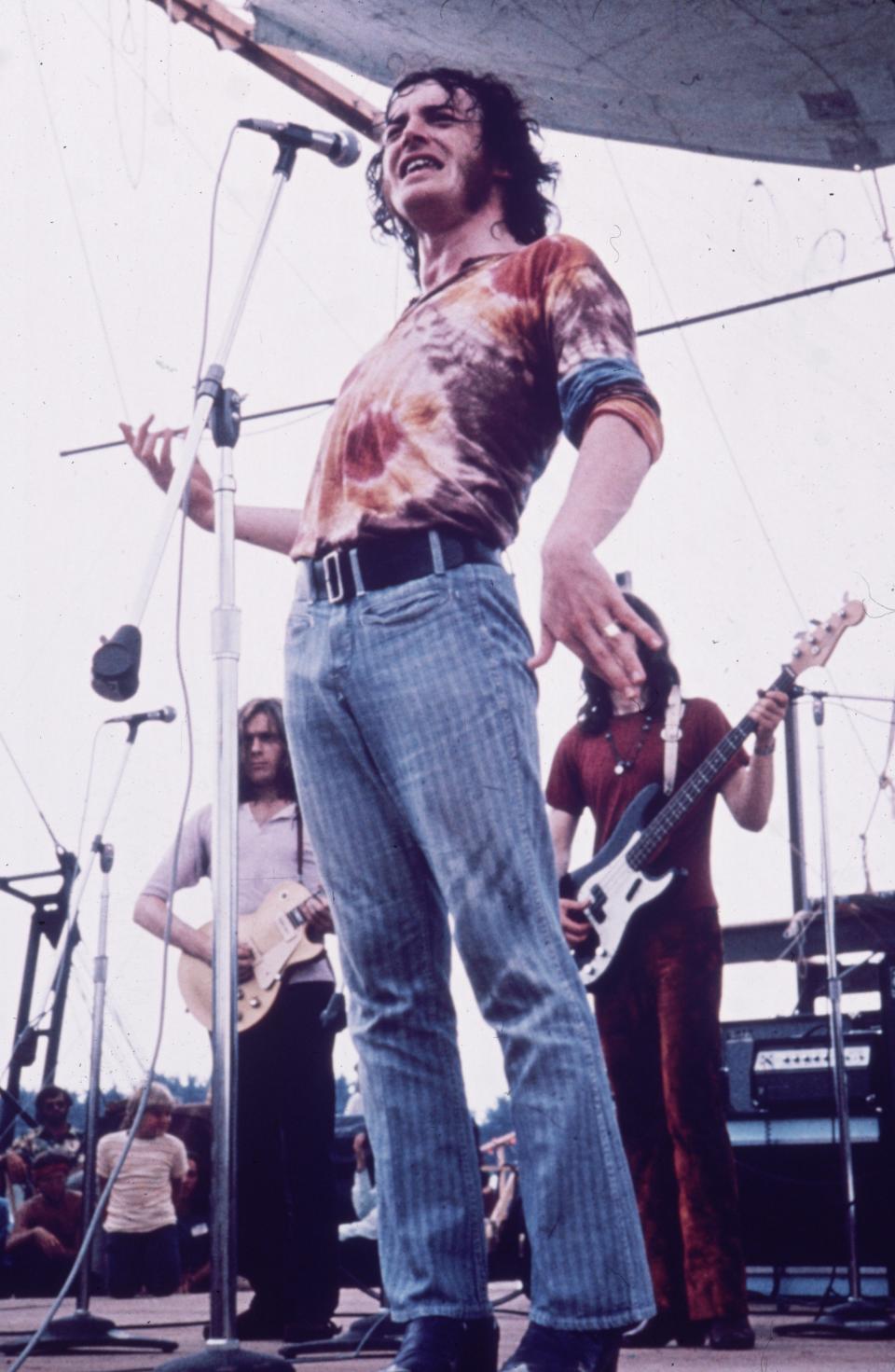 Joe Cocker performs on stage at Woodstock, wearing a tie-dye shirt and striped pants, with his backing band playing behind him