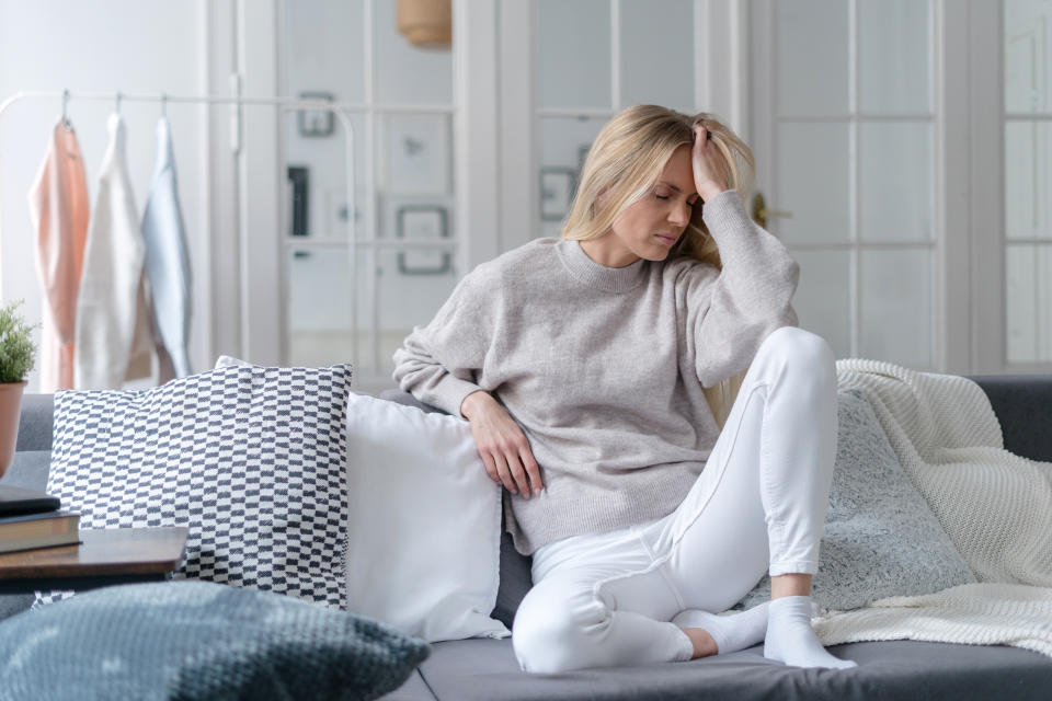 upset female suffering from migraine, sitting on sofa with pillows in living room and holding forehead, lonely tired or exhausted girl at home