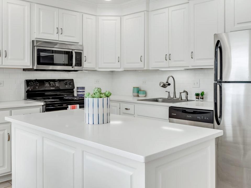 An all-white kitchen with stainless steel fridge, a decorative canister with artichokes in it, and black stovetop range