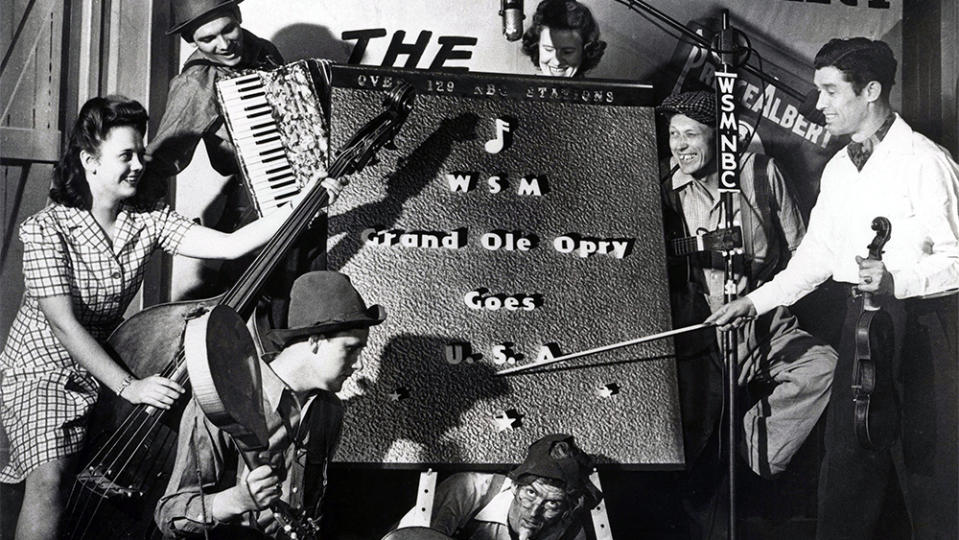 Clockwise from top of sign, Rachel Veach, Pete Ruby, Lonnie “Pap” Wilson, Jess Easterday, Thelma Smith in front of “WSM Grand Ole Opry Goes USA” sign, celebrating GOO joining the nation-wide NBC network, with sponsor Prince Albert sign in the background. Roy Acuff on right points to USA with his fiddle bow. 1943. - Credit: Courtesy of Grand Ole Opry Collection