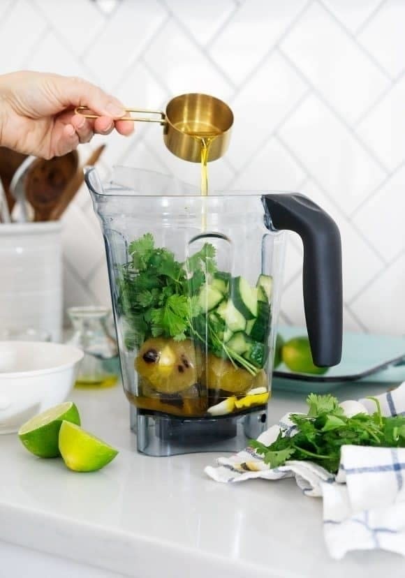 A person pours liquid from a brass measuring cup into a blender filled with fresh green vegetables and pears. Nearby, lime halves and herbs are on the counter