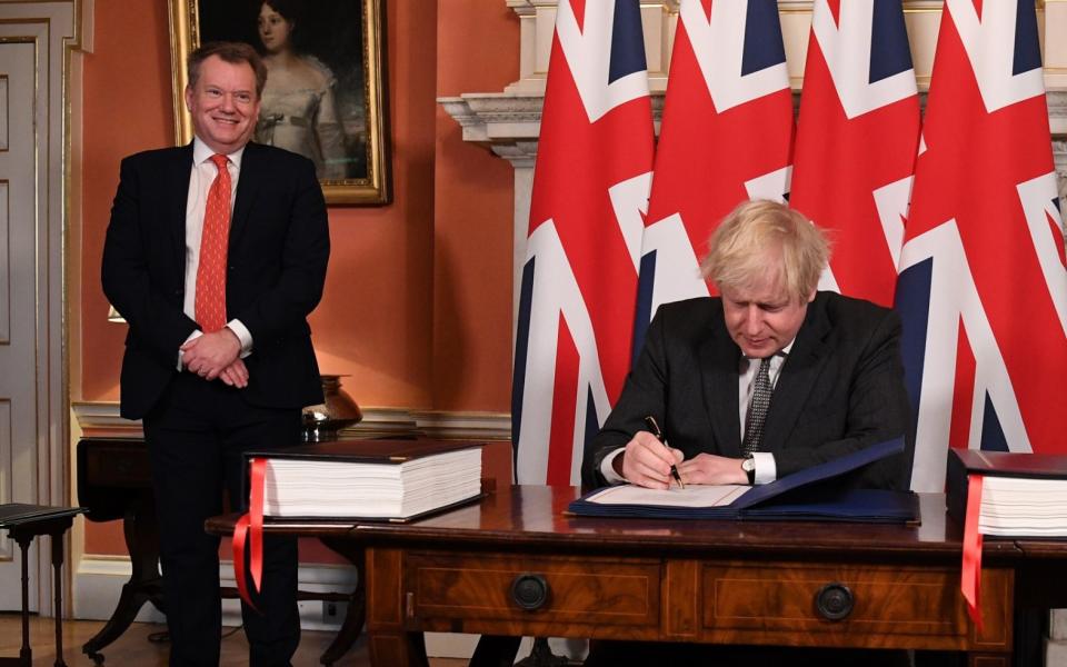 Boris Johnson sits at a desk signing, with Union flags hanging behind him. Lord Frost looks on, smiling