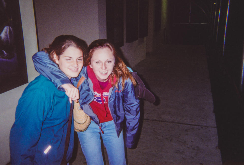 Two friends, one with brown hair and one with red hair, smile and pose with arms around each other. Both wear casual clothes including jeans and jackets