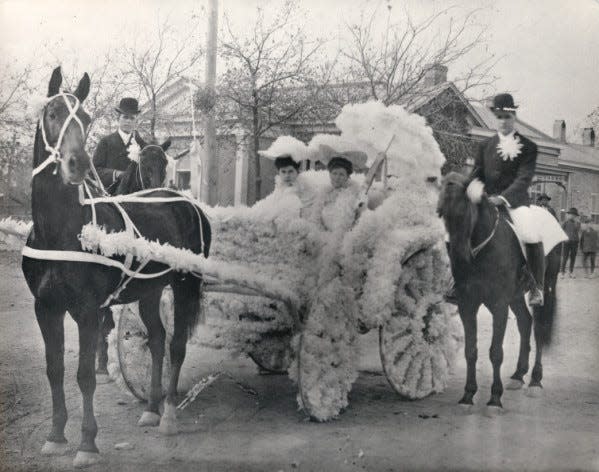 Ima Hogg and an unidentified woman are seated in a horse-drawn carriage, decorated with flowers for the Tekram parade, part of Houston's No-Tsu-Oh festival. Two unidentified men sit atop horses on each side of the carriage. Date late 1890s