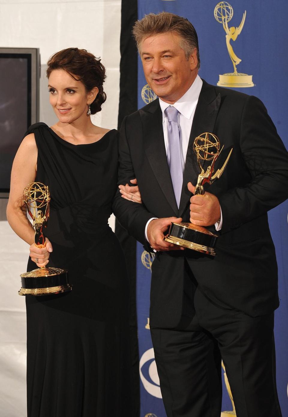 tina fey and alec baldwin each hold an emmy award and pose for photos while standing in front of a blue backdrop, she wears a black sleeveless dress, he wears a black suit with a white collared shirt and lavender tie