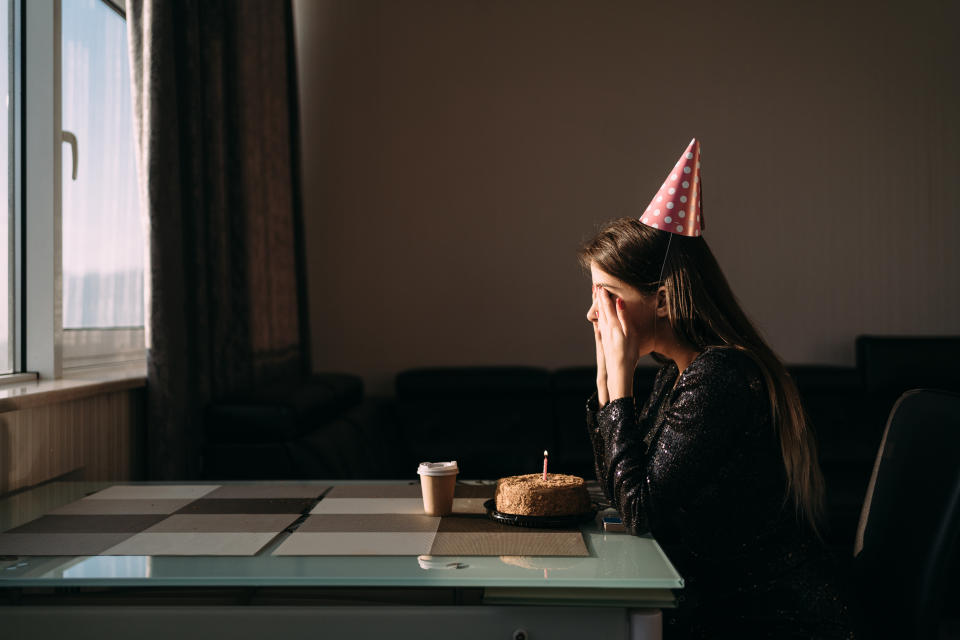 A woman sits alone at a table with a small cake and coffee, wearing a party hat and black outfit. She appears to be celebrating her birthday