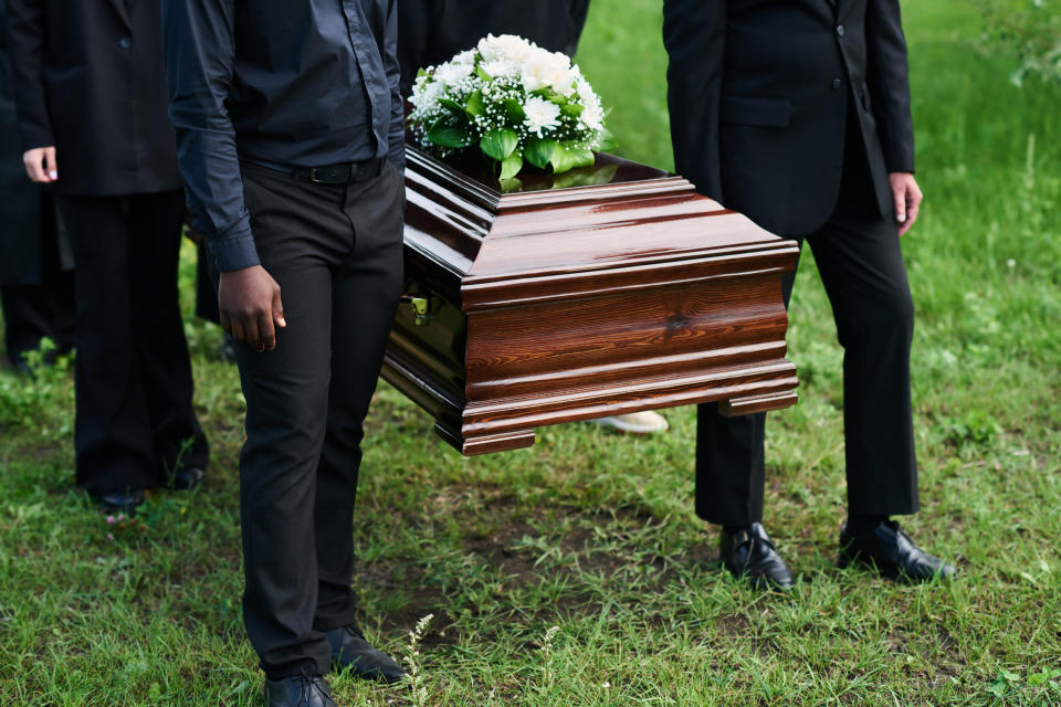 Several individuals in formal black attire carry a wooden casket with a flower arrangement