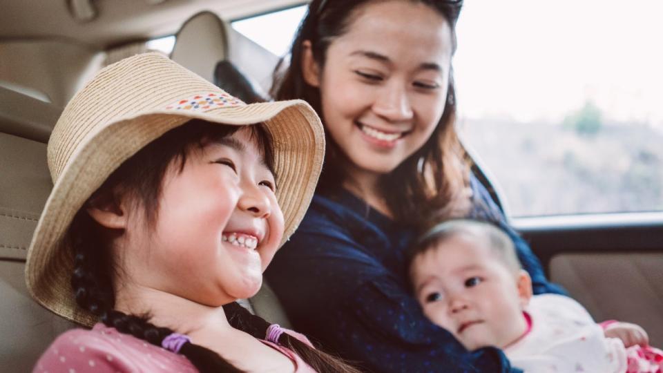 Asian woman sits in the back seat of a car with her infant child in her arms. Her daughter sits next to her, smiling and wearing a floppy straw hat.