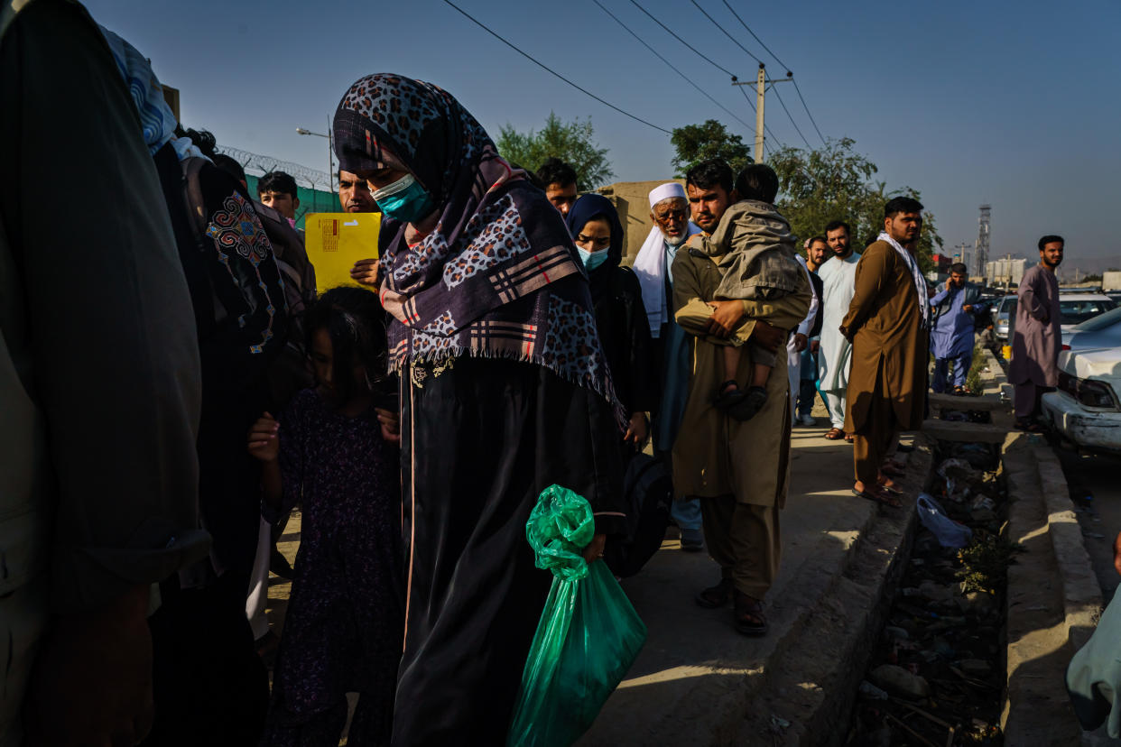 KABUL, AFGHANISTAN -- AUGUST 19, 2021: Afghans make their way the road to the military entrance of the airport for evacuations, in Kabul, Afghanistan, Thursday, Aug. 19, 2021. Here Afghans are made to wait behind the first Taliban checkpoint, before bringing their worldly possessions one can only carry in both hands to the military gate for evacuations out of the country. A few hundred people congregate there listening to spotty announcements from a skinny Taliban fighter with neck-length hair standing on an elevated platform, wearing a surgical mask for covid19 prevention, but barely covering his mouth. On his right hand a cold water bottle. On his left hand a semi-automatic pistol and a walkie talkie radio that he occasionally waves to the crowd to get their attention. The Taliban fighter with the pistol announces that they will call out names of countries and people with approval for flights to those countries will be allowed to pass. (MARCUS YAM / LOS ANGELES TIMES)