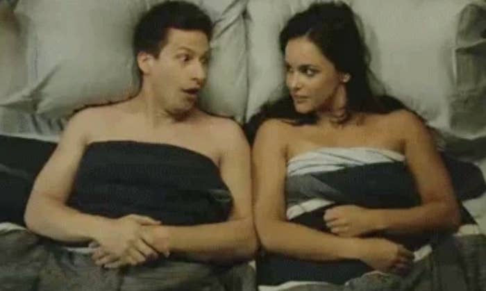 Andy Samberg and Melissa Fumero, wrapped in blankets, lie on a bed looking at each other with surprised expressions