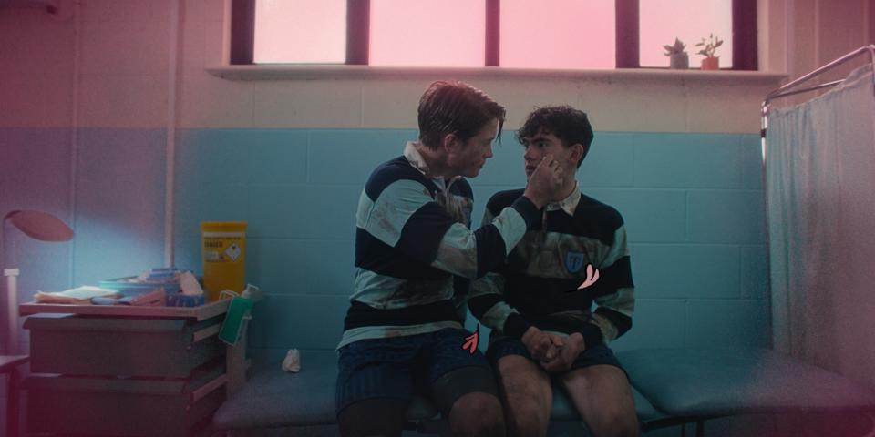 Kit Conner and Joe Locke sit close to each other in a nurse's office in Heartstopper. They wear rugby uniforms.