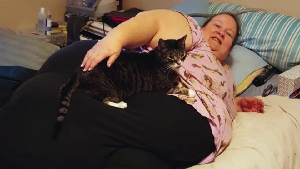 Janine lays on her bed in a pink shirt with a bird pattern and black pants, while her cat lays on top of her.
