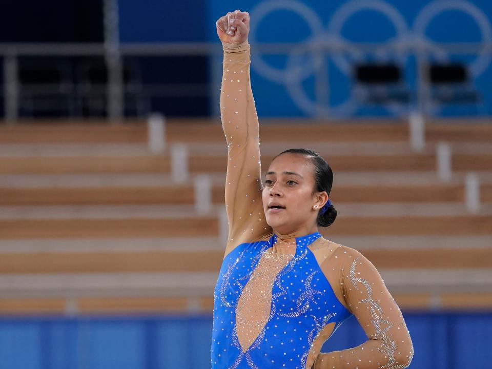 Gymnast in a blue leotard raising her fist in the air and kneeling on the ground after her routine in the Tokyo Olympics.