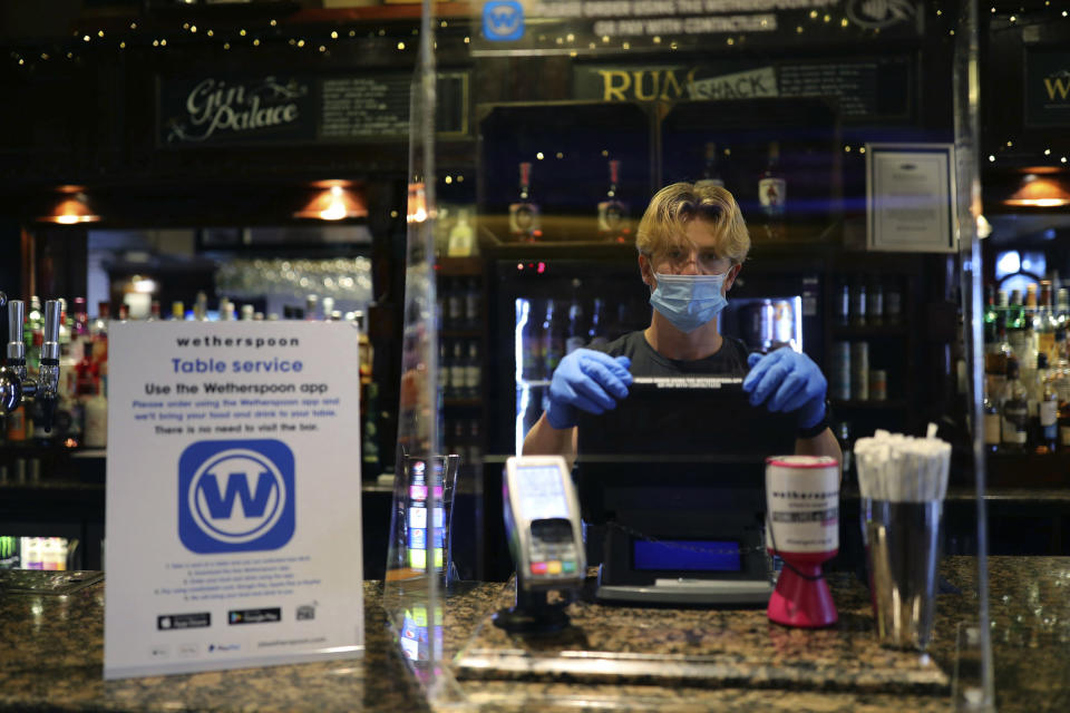 A member of staff wearing PPE, stands behind a screen as he waits to serve drinkers at the reopening The Toll Gate, a Wetherspoons pub in Hornsey, north London, as coronavirus lockdown restrictions are eased across the country, Saturday July 4, 2020. Restrictions which were imposed on March 23 have been eased allowing businesses including pubs, restaurants and hair salons, to reopen to members of the public with measures in place to prevent the spread of the coronavirus. (Aaron Chown/PA via AP)