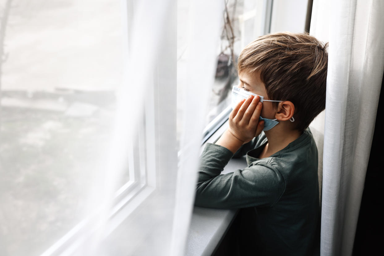 A child wearing a face mask looks out of a window with their elbows resting on the window sill and chin resting on their hands.