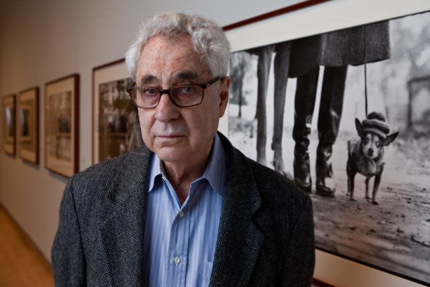 Elliott Erwitt, wearing a gray coat and blue shirt, poses in front of his photo of a small leashed dog in a hat