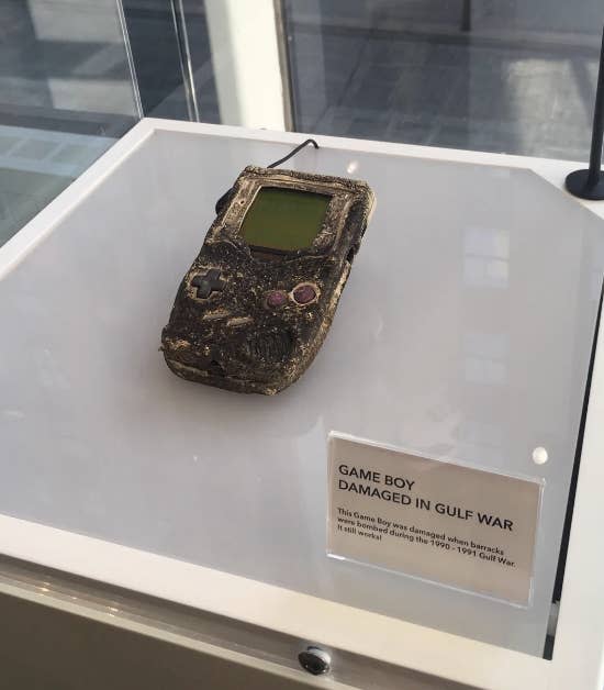 Damaged Nintendo Game Boy on display, labeled "Game Boy damaged in Gulf War." The case shows severe wear, and it appears burnt. Accompanying text explains its history