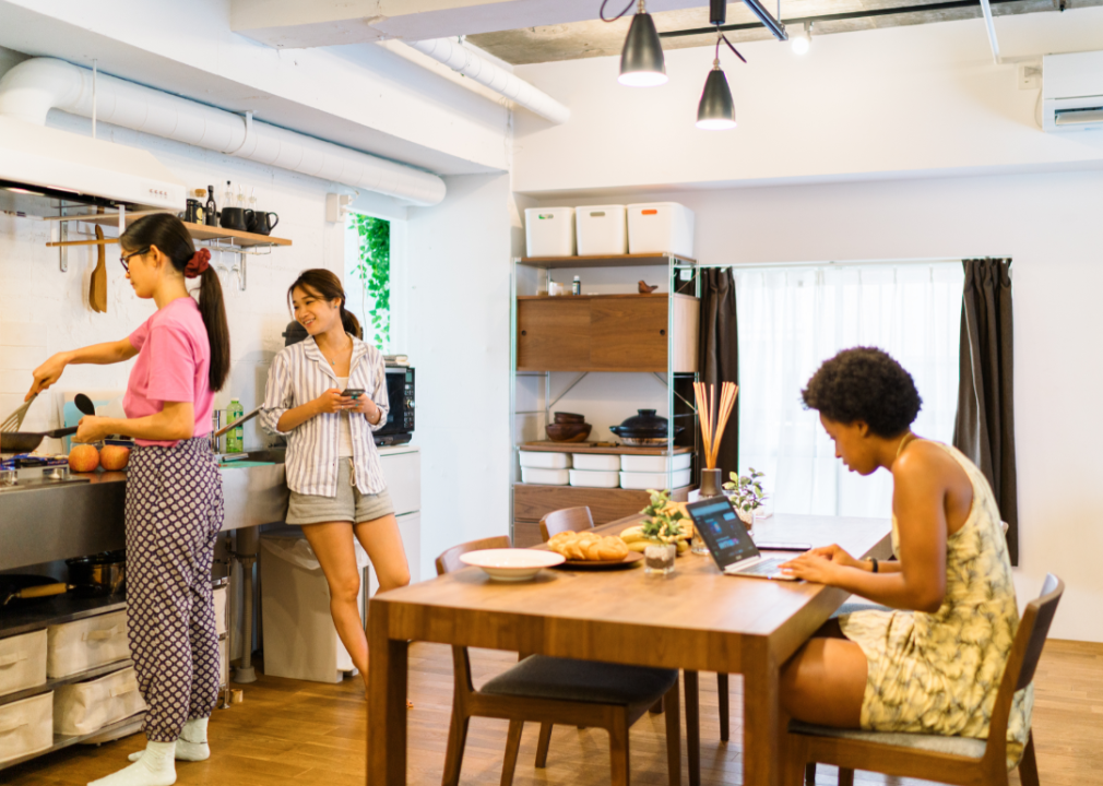 Three roommates share the kitchen, two cooking and one using a laptop at the table.