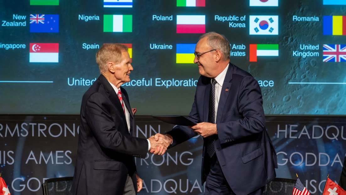  Two white men in dark suits shake hands in front of a backdrop of many flags. 