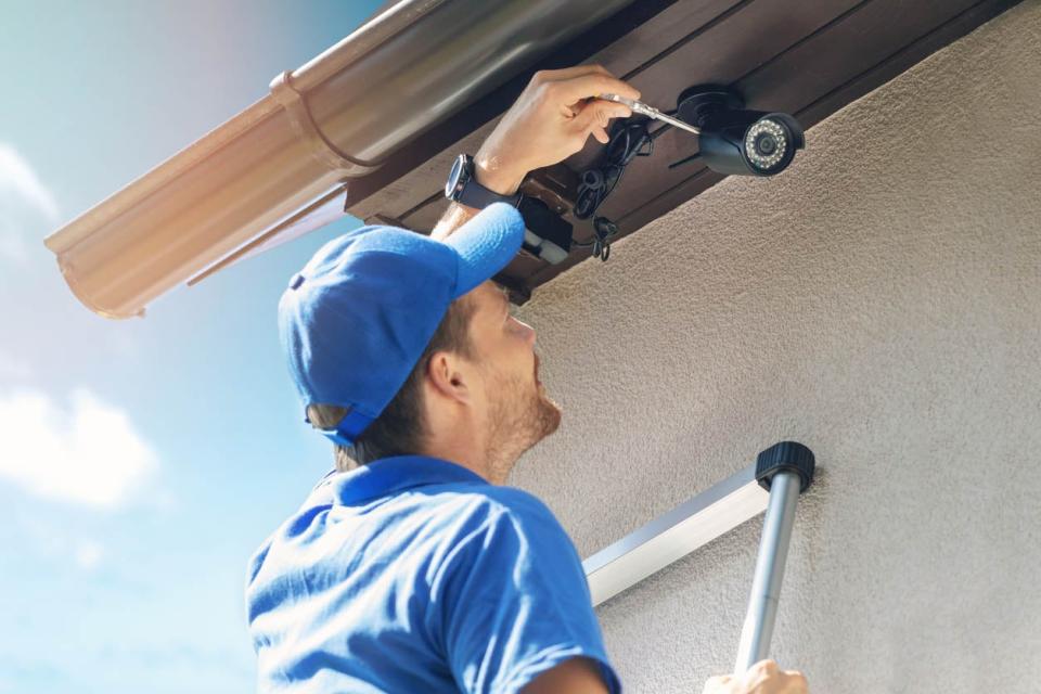 A worker in a blue shirt and blue hat is on a ladder fixing a security camera.