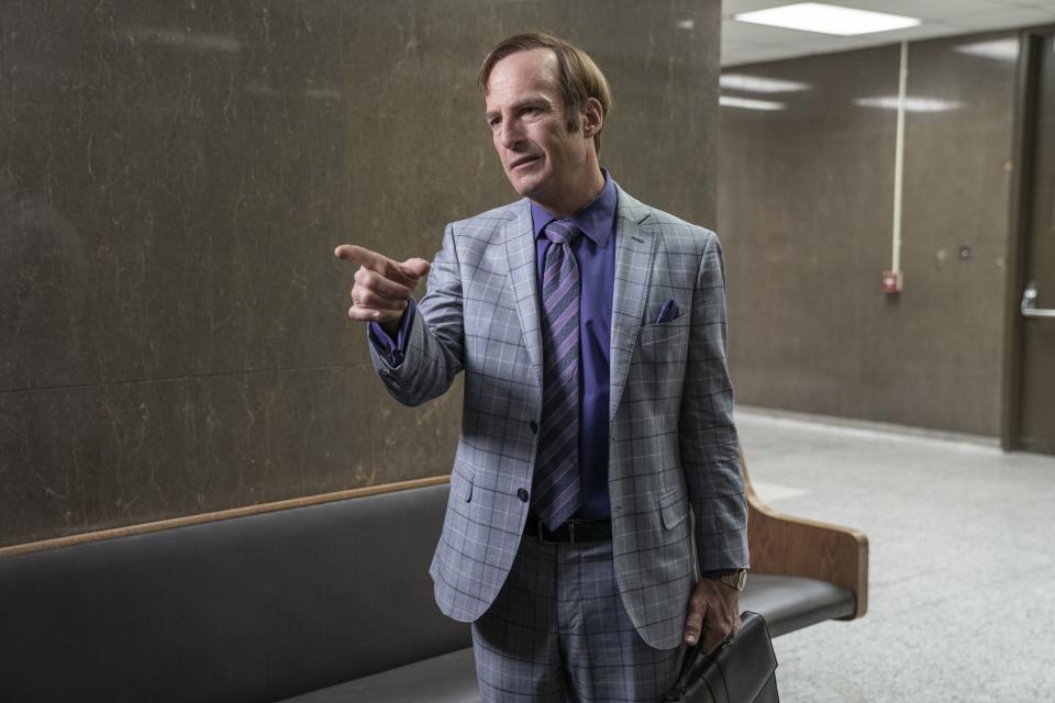 Man in a checked suit with tie pointing, a briefcase in hand, standing in a hallway