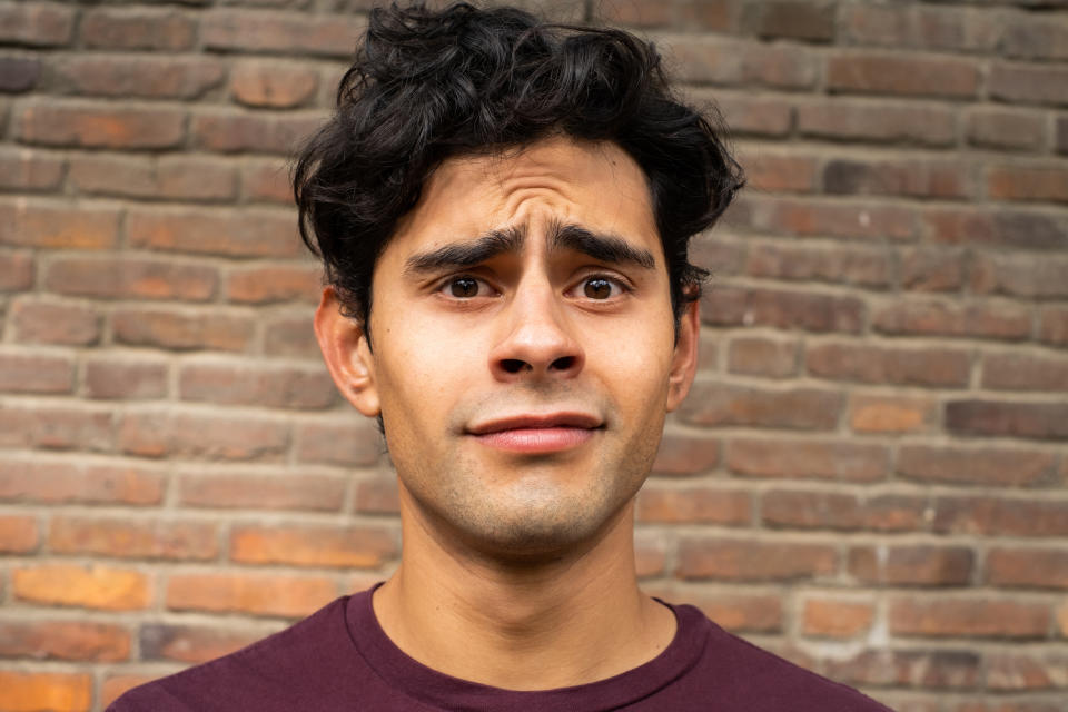 A person with a puzzled expression stands in front of a brick wall, wearing a plain shirt