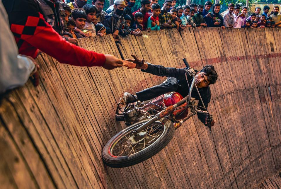 A man riding a motorcycle on the "Well Of Death" grabs a tip offered by an audience member as he rides past, vertically to the ground.