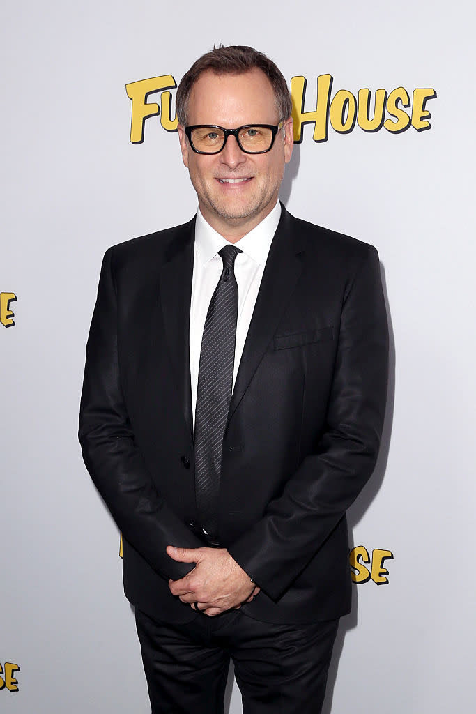 Dave Coulier standing in front of a 'Fuller House' sign, wearing a classic black suit and tie