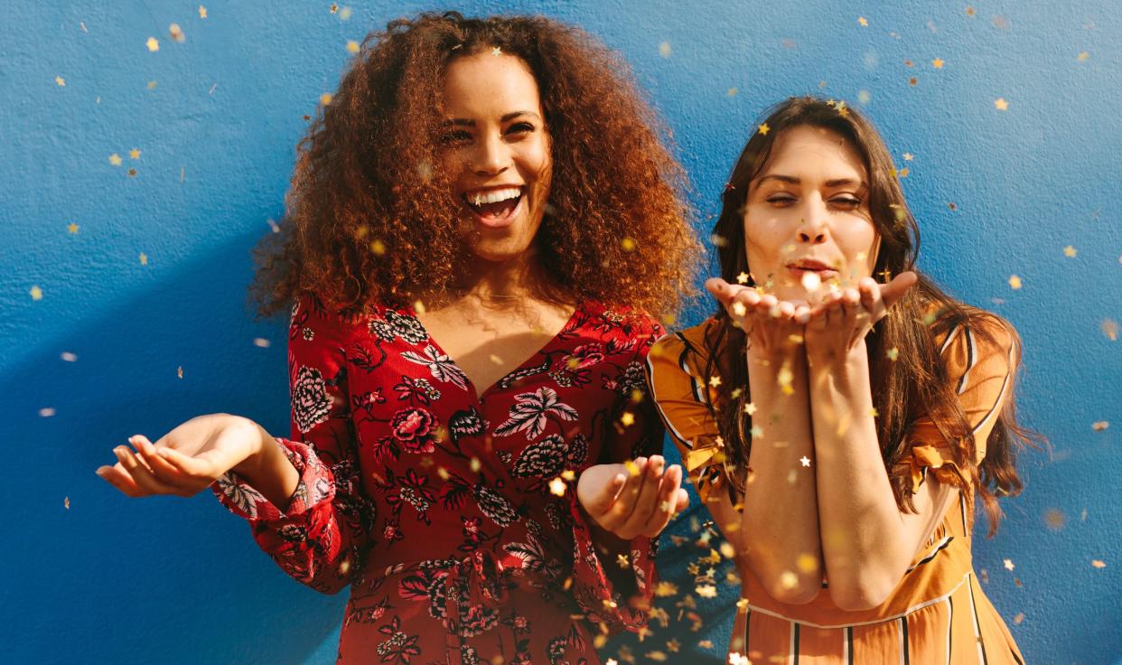 Attractive young women blowing glitters against blue background. Mixed race female friends having fun with glitters.