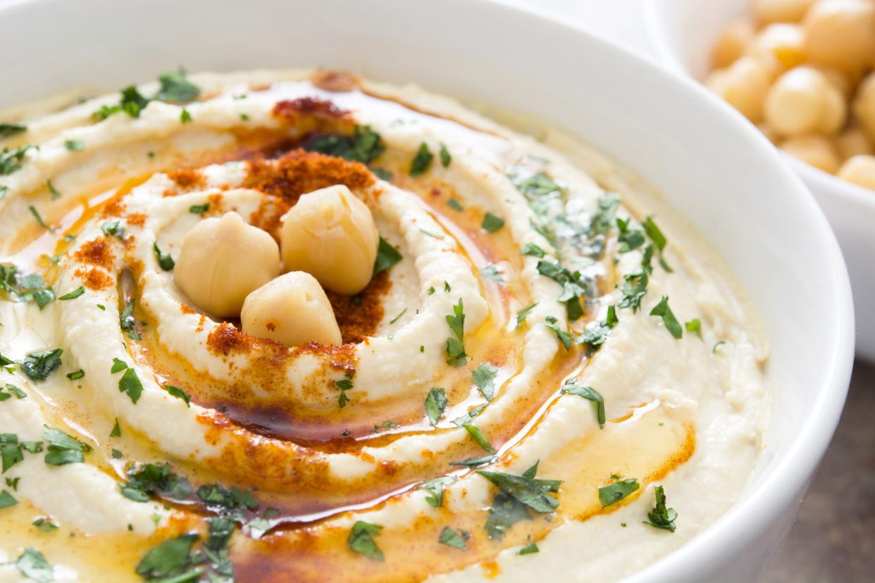 Closeup of hummus with oil and herbs in a white bowl with a bowl of chickpeas blurred in the background