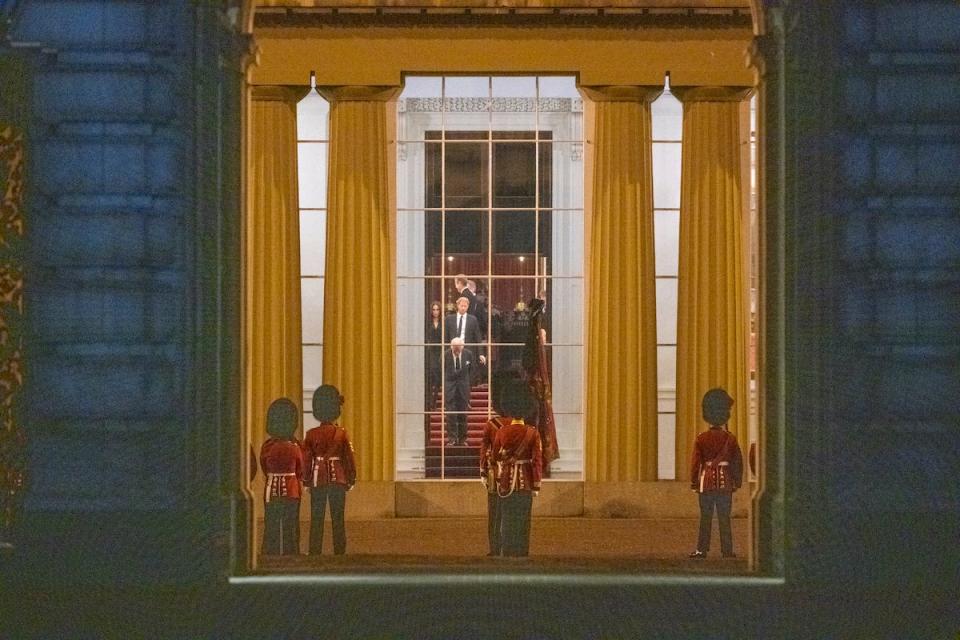 A photo shows King Charles III, Prince Harry, and Meghan Markle at Buckingham Palace through a window, while royal soldiers stand outside.