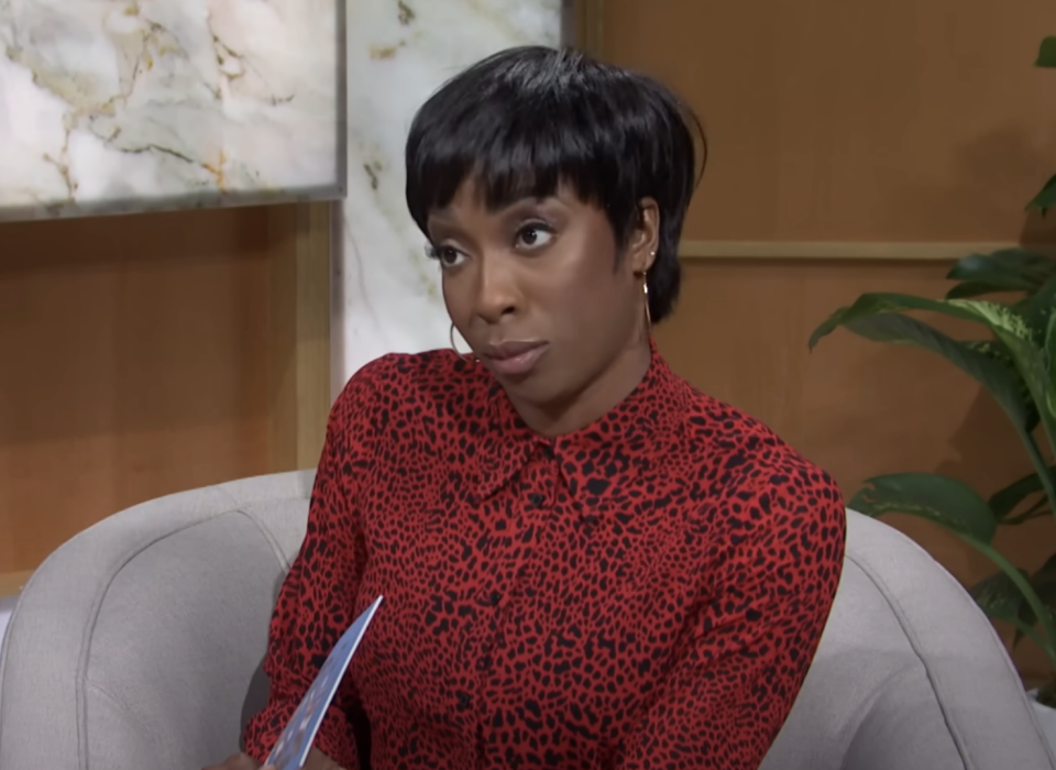 A person dressed in a leopard-print blouse is seated on a sofa, looking at someone off-camera with a serious expression, holding a piece of paper