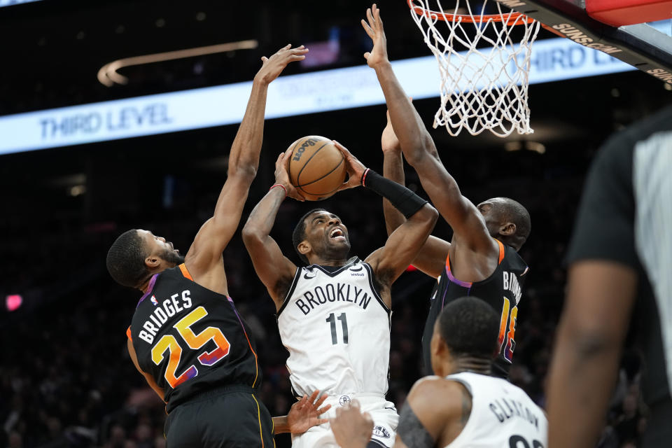 Brooklyn Nets guard Kyrie Irving (11) shoots between Phoenix Suns forward Mikal Bridges (25) and center Bismack Biyombo during the first half of an NBA basketball game, Thursday, Jan. 19, 2023, in Phoenix. (AP Photo/Matt York)