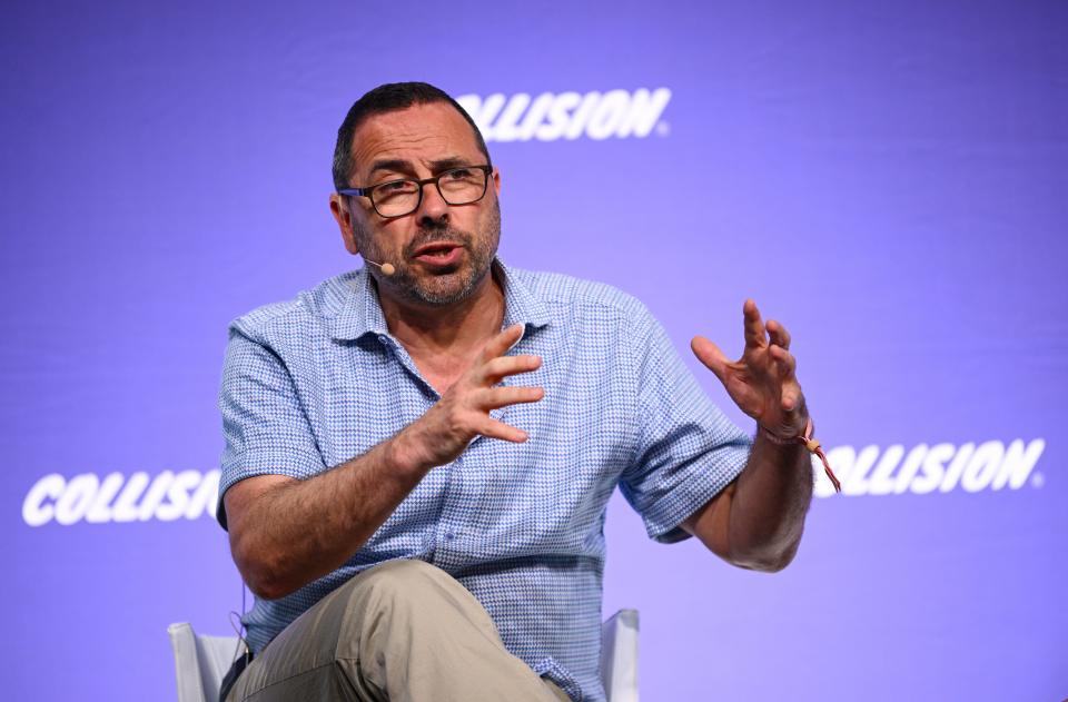 Andrew Feldman sits on a stage in casual clothes speaking and gesticulating wearing a head mic in front of a purple background that say "Collision."