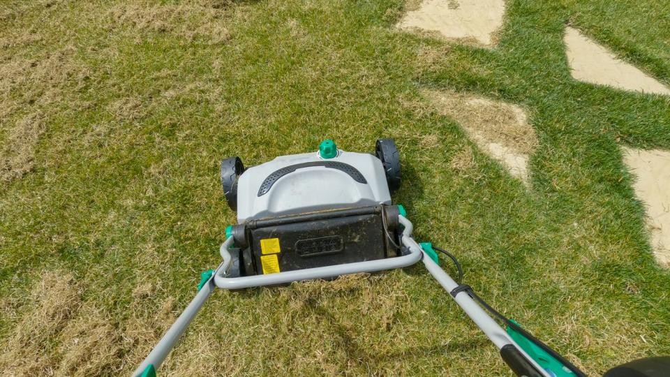  Gardener using grass aerator while taking care of backyard lawn. First person view of spring home garden work for green lawn growth enhancement. Practical machinery for landscaping.