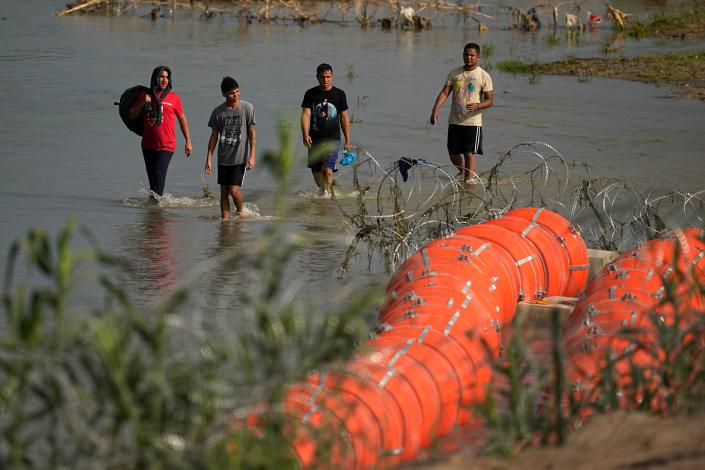 Migrants walk past the site where workers are assembling large buoys to be used as a border barrier along the banks of the Rio Grande in Eagle Pass, Texas, Tuesday, July 11, 2023. The floating barrier is deployed in an effort to prevent migrants from entering Texas from Mexico.