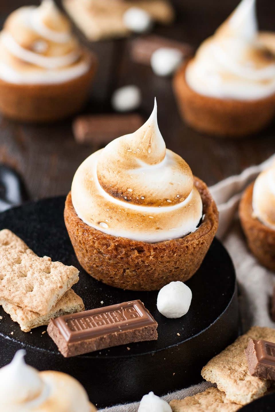 s'mores cookie cups with a cookie cup crust, chocolate filling, and toasted meringue topping, on a plate with graham crackers, hershey's chocolate square, and marshmallow