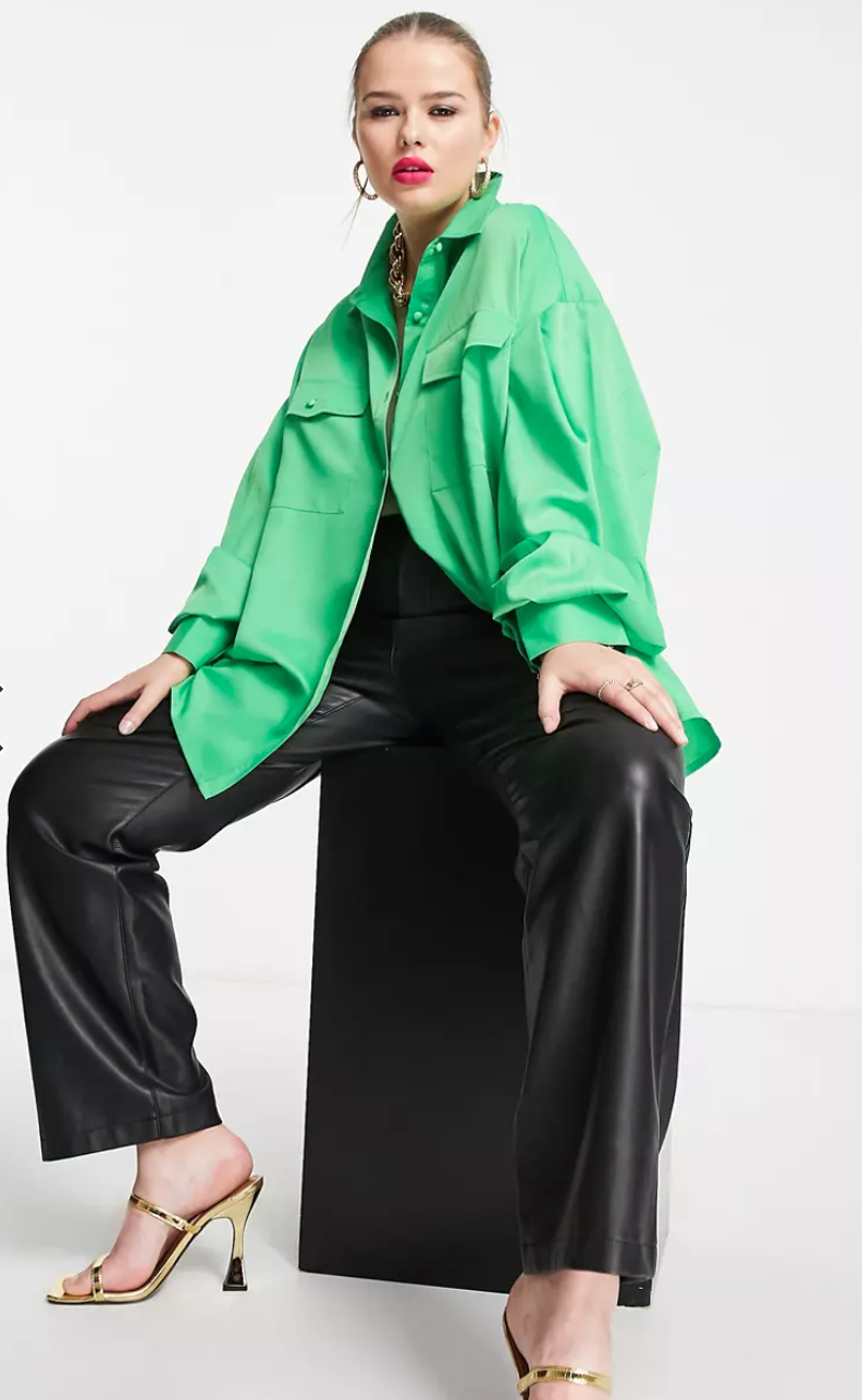 Model with hair tied back sits on a stool wearing a bright green Extro & Vert Plus pleated oversized shirt over shiny dark trousers and high gold sandals.