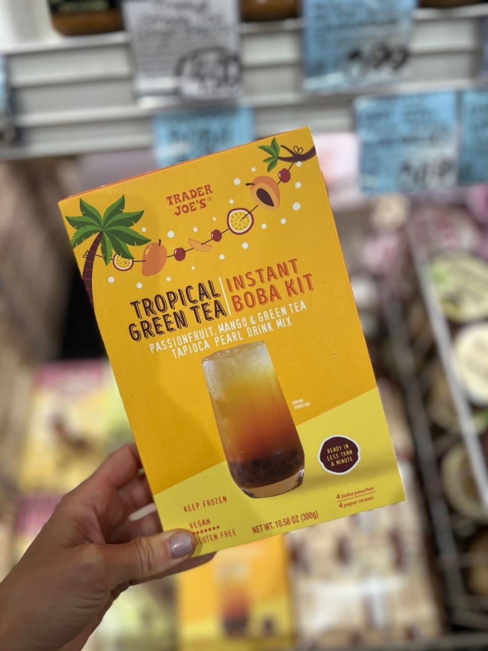 Hand holding a Trader Joe's Tropical Instant Green Tea Boba Kit box. Box text includes "Passionfruit, Mango, & Green Tea Tapioca Pearl Drink Mix."