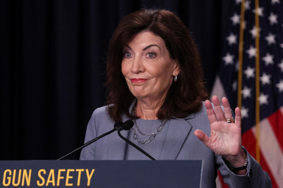 Kathy Hochul holds up her left hand, palm out, in front of an American flag at a podium labeled with: Gun safety.
