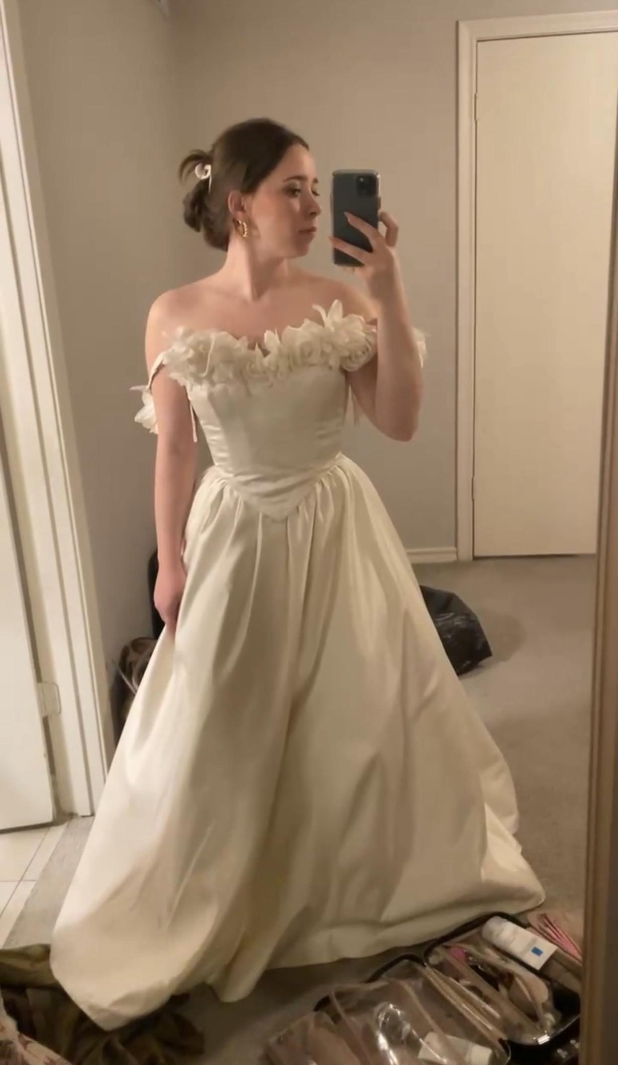 A woman taking a mirror selfie while wearing an off-the-shoulder wedding dress with floral rosettes on the neckline and a V-shaped waist.