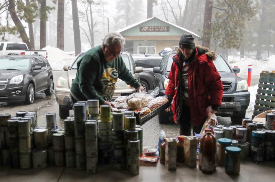 Two people stand outside and near a table loaded with canned food while snow is visible on the ground and houses behind them.
