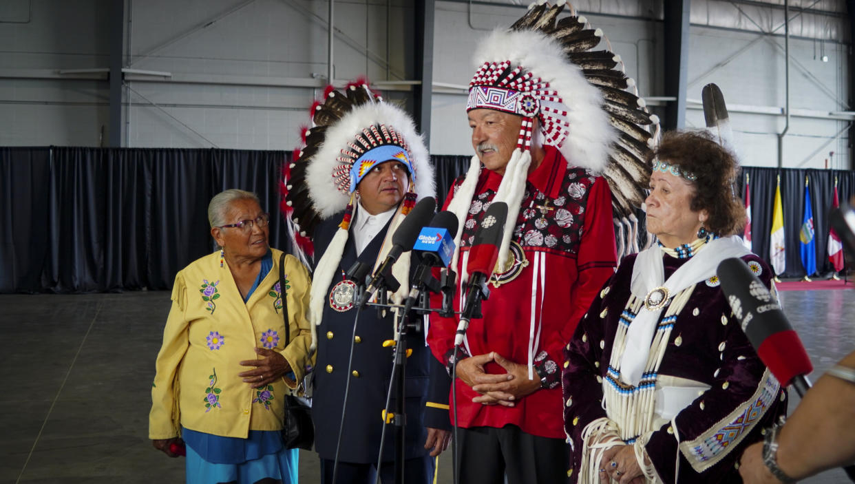 Four people, two wearing feather headdresses, stand in a large building before microphones.
