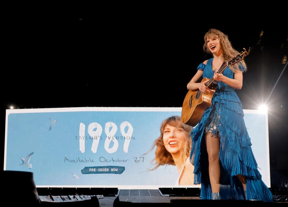Taylor onstage holding a guitar with an ad for "1989 (Taylor's Version)" behind her
