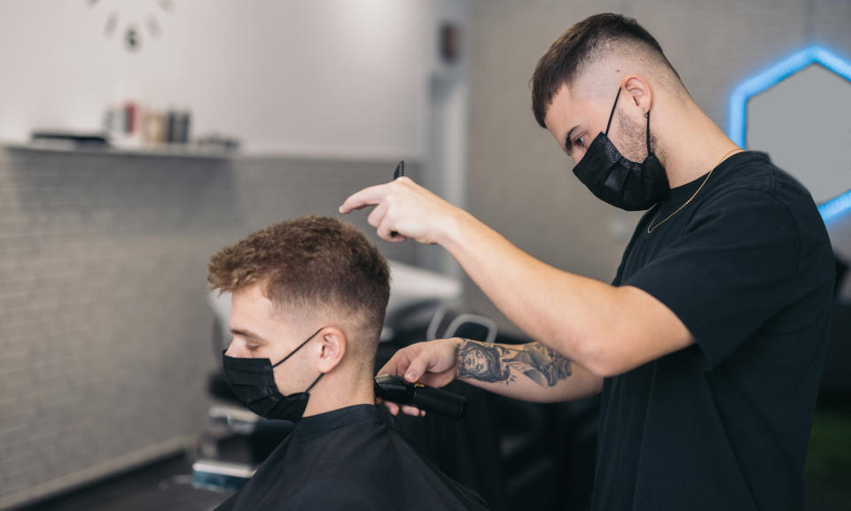 A barber wearing a surgical mask works on a young man who's also wearing a mask