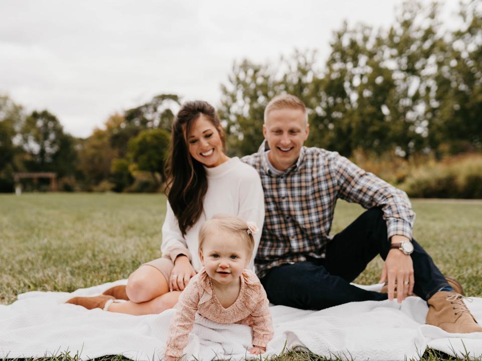 Brennan and Erin Schlagbaum sitting on a picnic blanket while watching their young daughter crawl across a grassy lawn.