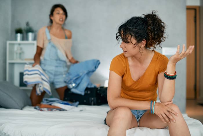 Two women arguing in a bedroom, one sitting on a bed with a frustrated expression, the other standing holding clothes