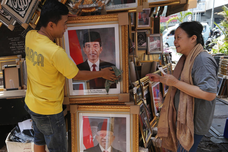 In this Wednesday. Oct. 16, 2019, photo, a vendor talks to a customer as he cleans a portrait of Indonesian President Joko Widodo and Vice President-elect Ma'ruf Amin, bottom, displayed at his stall in Jakarta, Indonesia. Known for his down-to-earth style with a reputation for clean governance, Widodo's signature policy has been improving Indonesia's inadequate infrastructure and reducing poverty, which afflicts close to a tenth of Indonesia's nearly 270 million people. But raising money would be harder at a time of global economic slowdown, major trade conflicts and falling exports. (AP Photo/Tatan Syuflana)