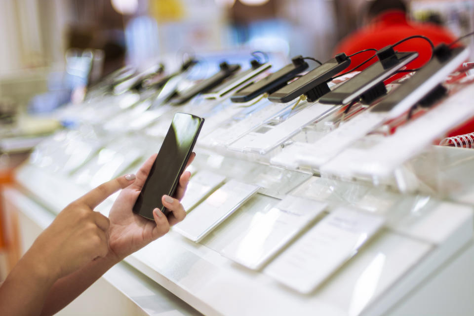 A person is holding a smartphone while their finger hovers over the screen. Multiple smartphones are displayed in the background in a store setting