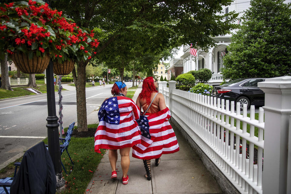 Parade goers draped in American flags walk down the street before a Fourth of July parade begins Saturday, July 4, 2020, in Bristol, R.I. The town, which lays claim to the nation's oldest Independence Day celebration in the country, held a vehicle-only scaled down version of its annual parade Saturday due to the coronavirus pandemic. (AP Photo/David Goldman)